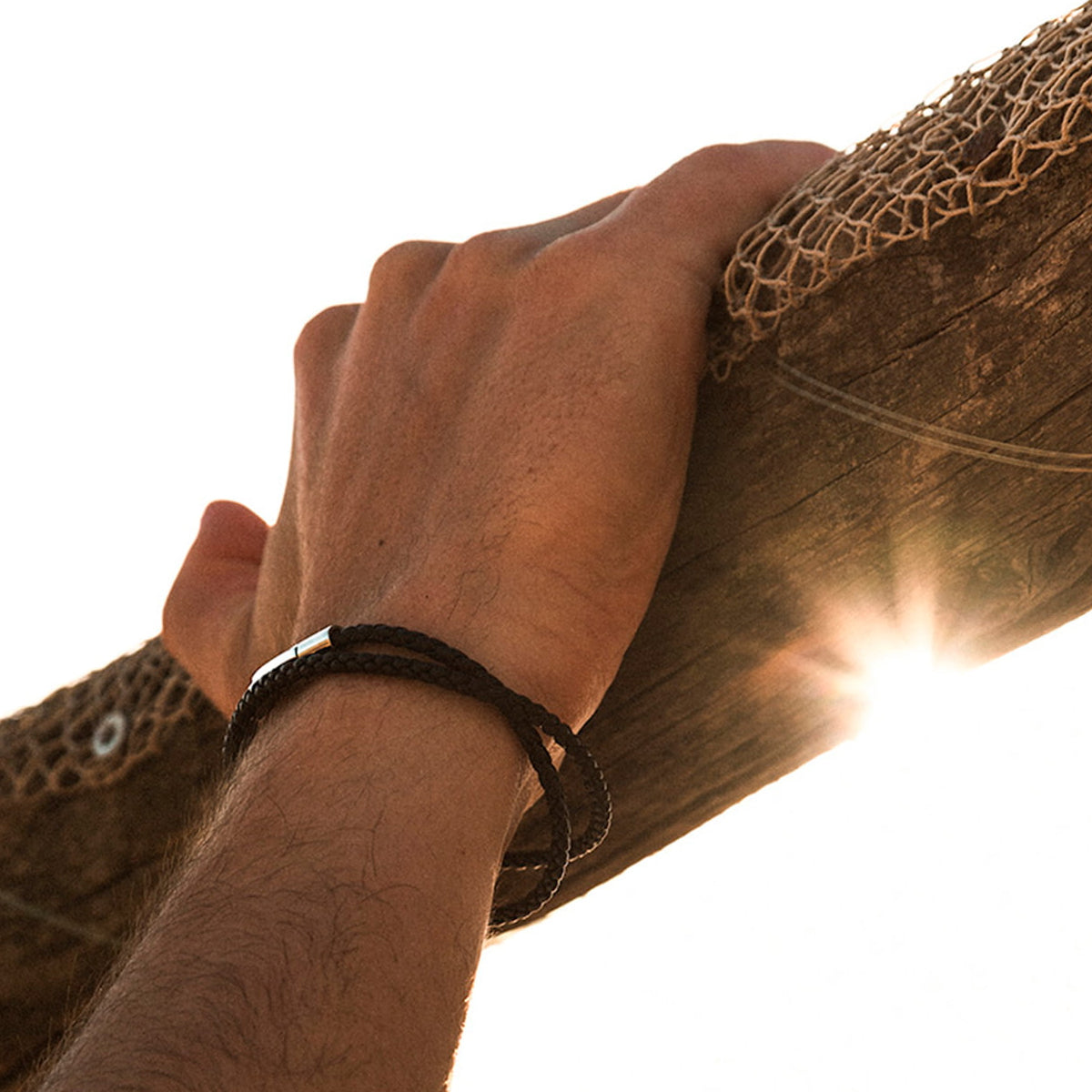 Close-up of a hand wearing a black braided bracelet holding onto an outdoor wooden structure with sunlight shining through.