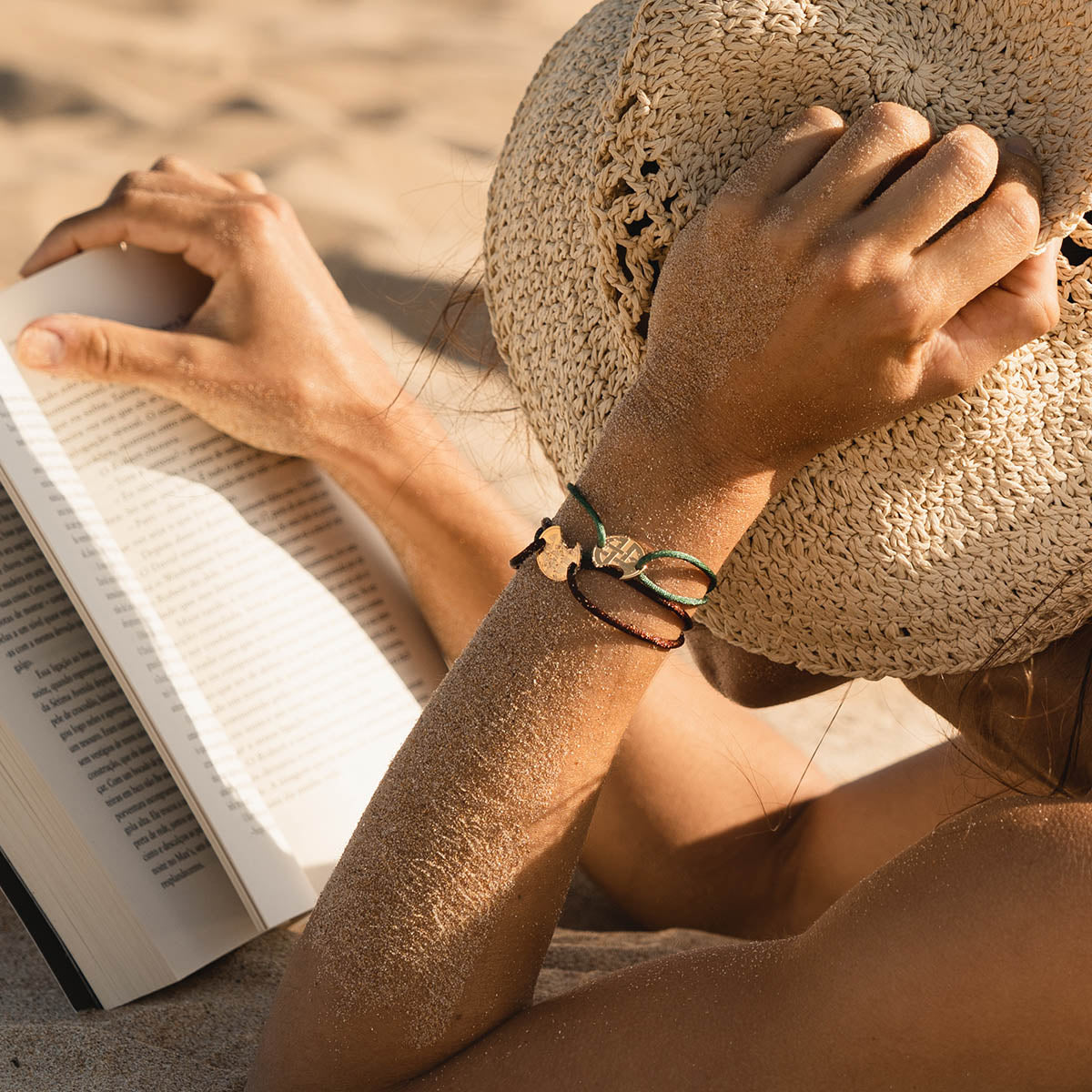 Woman on beach wearing AN JEWELS JEWELRY Mod. AV.BGLBGGGR bracelet, reading book and holding straw hat.